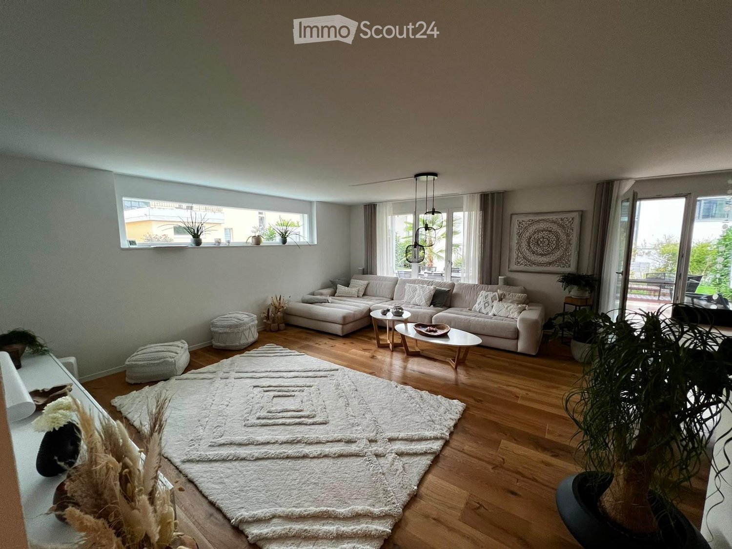 Modern living room with light-colored sofa, coffee table, ottoman, large white rug, and potted plants. Large windows and hanging pendant lights.