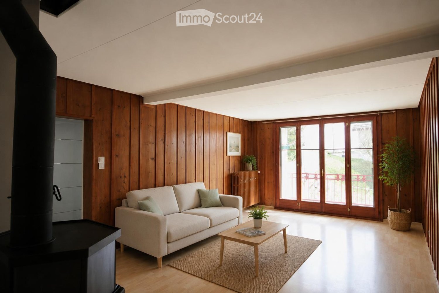 Modern living room with wooden walls and floor, large sliding glass doors leading to a balcony, a light-colored sofa, a wooden coffee table, and decorative plants.