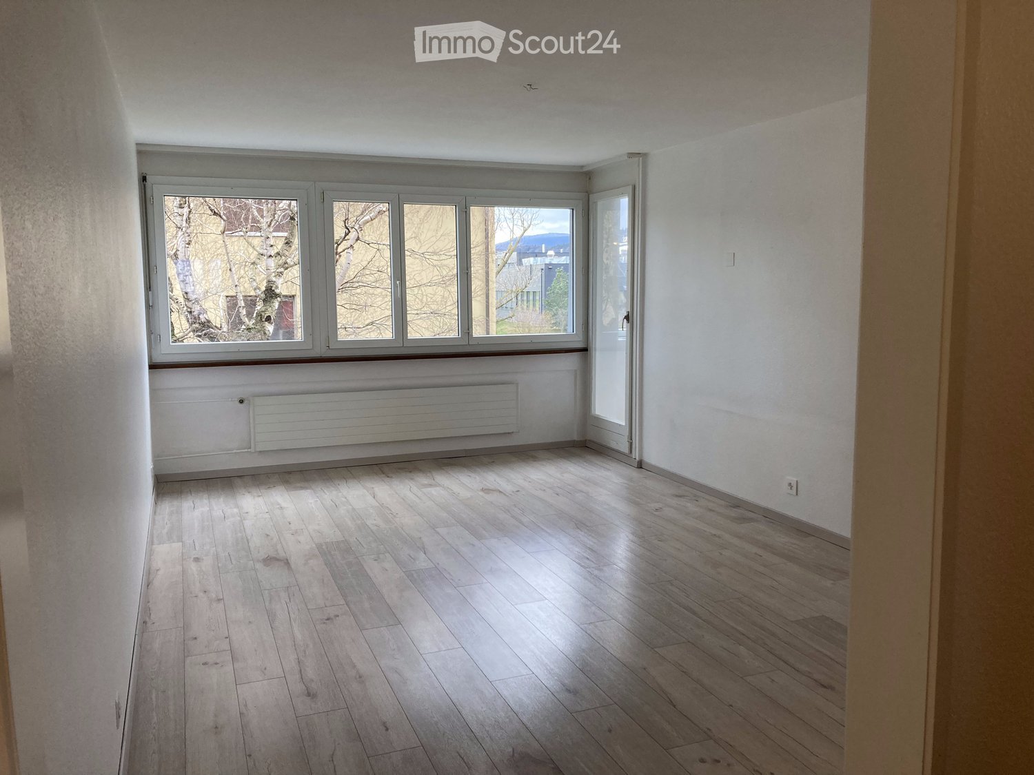 Empty room with light wooden flooring, white walls, large windows, and a radiator below the windows.