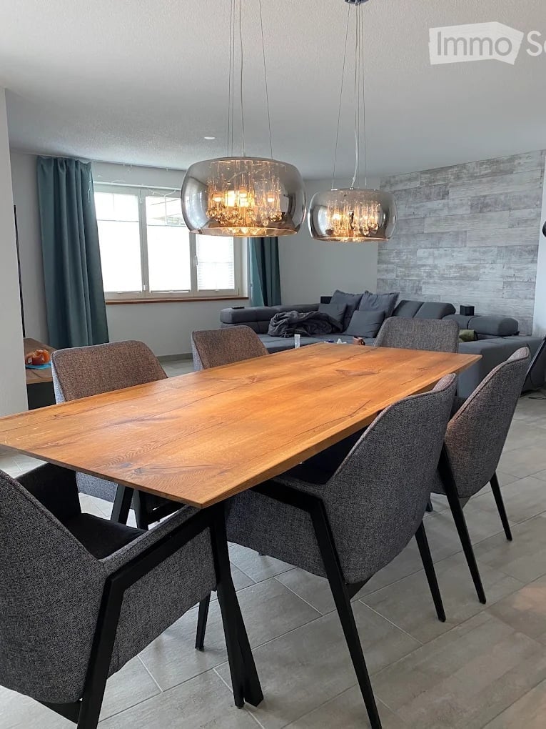 Modern dining area with a wooden table, six grey chairs, a grey sofa, and two pendant lights hanging from the ceiling.