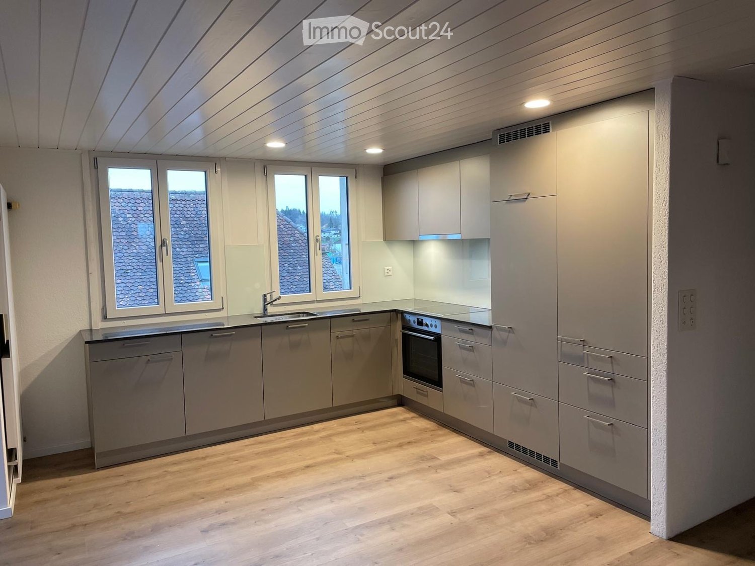 Modern kitchen with light grey cabinets, wooden flooring, and large windows offering a view of the roof.