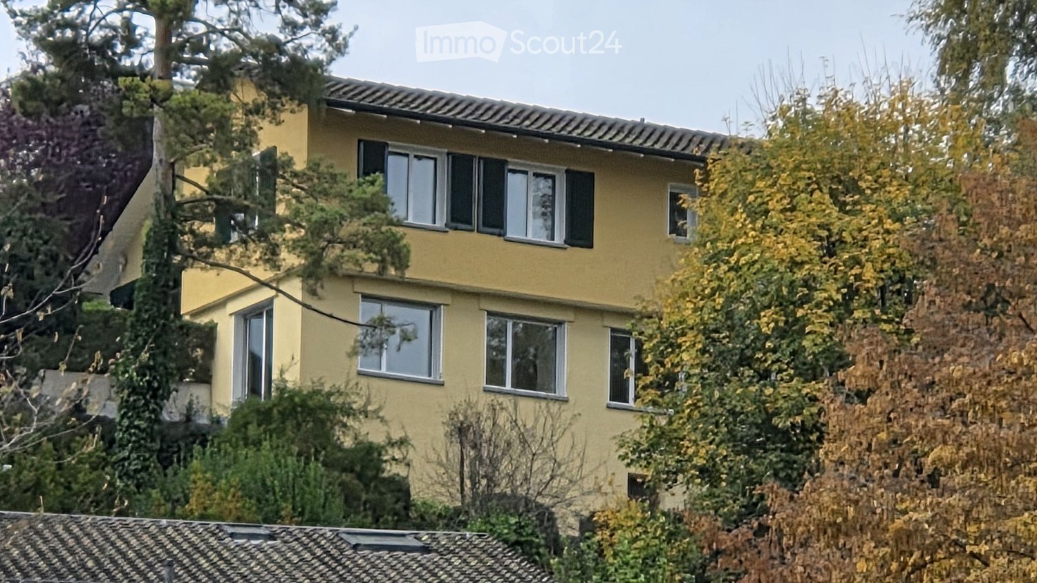 Two-story yellow house with multiple windows and a tiled roof, surrounded by trees and shrubs.