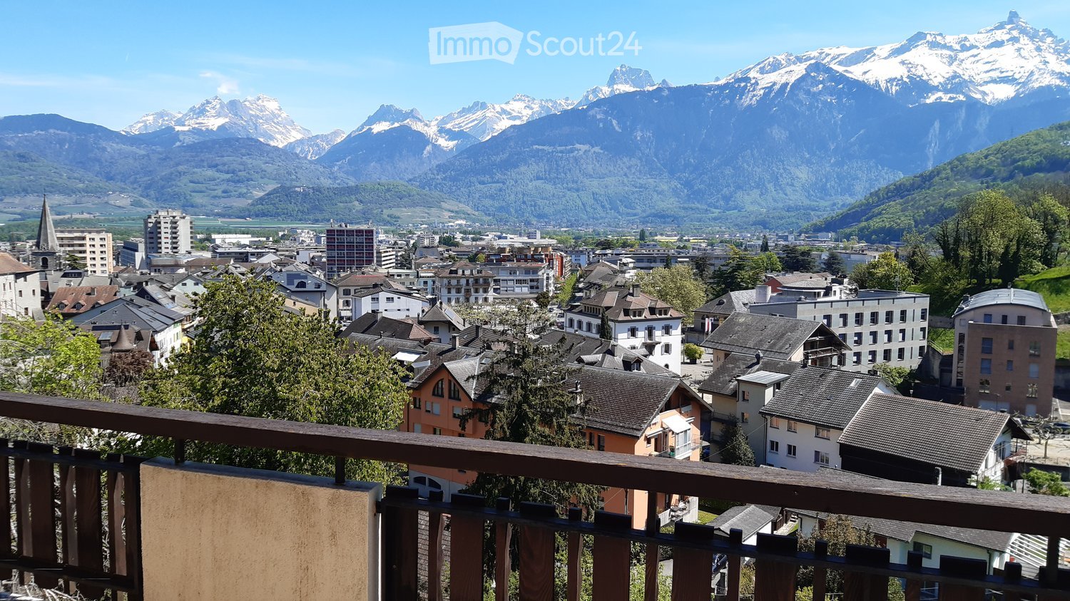 Panoramic view from a balcony overlooking a town with a mix of residential and commercial buildings, surrounded by lush greenery and snow-capped mountains in the background.