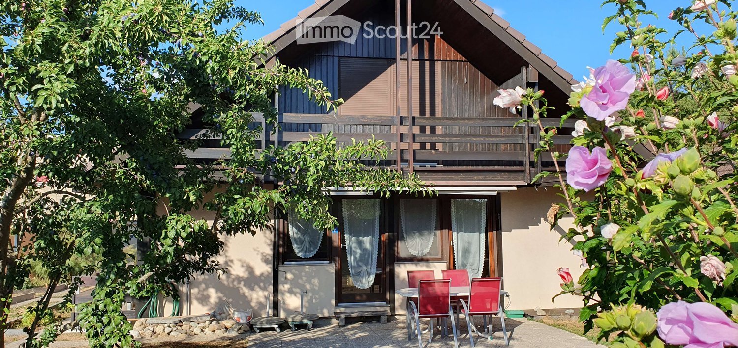 Two-story house with a wooden balcony, surrounded by greenery and flowers, with a small patio area featuring a table and chairs.