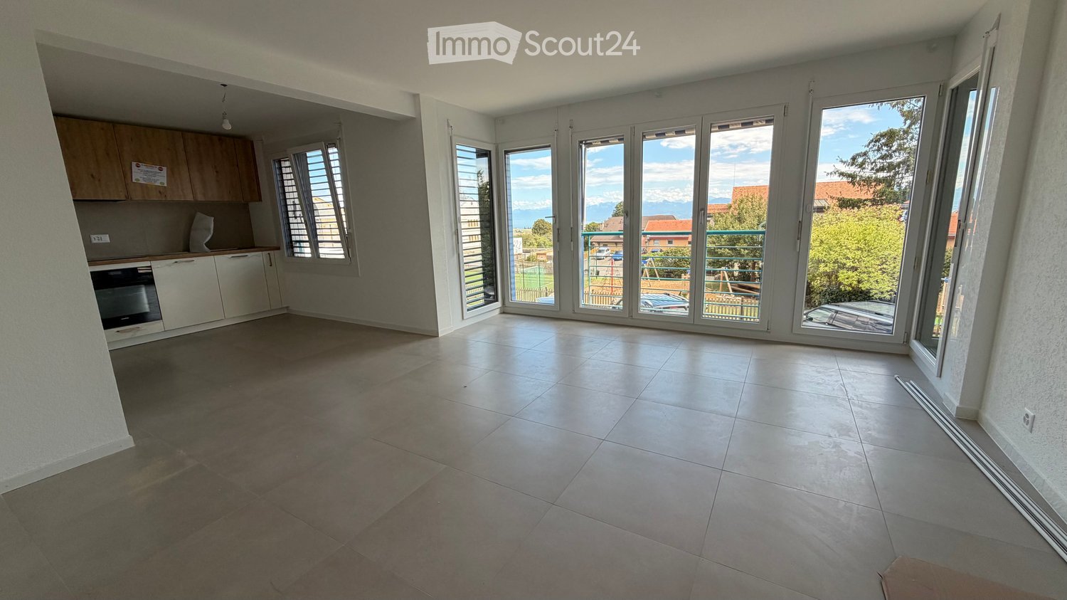 Empty room with tiled floor, large sliding glass doors leading to a balcony, and a small kitchen area with cabinets and appliances.