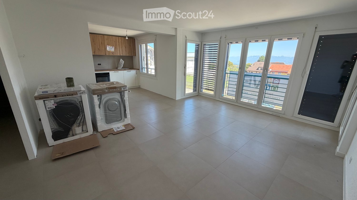Empty room with tiled floor, large windows, and a view of the sea. Kitchen area visible in the background with cabinets and appliances.