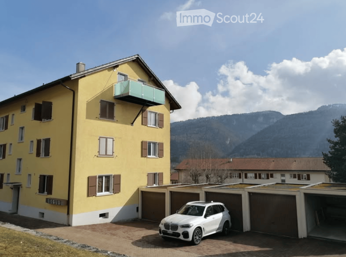 Yellow multi-story building with multiple windows and shutters, featuring a balcony and a garage area with a parked car. The background shows a scenic mountain view.