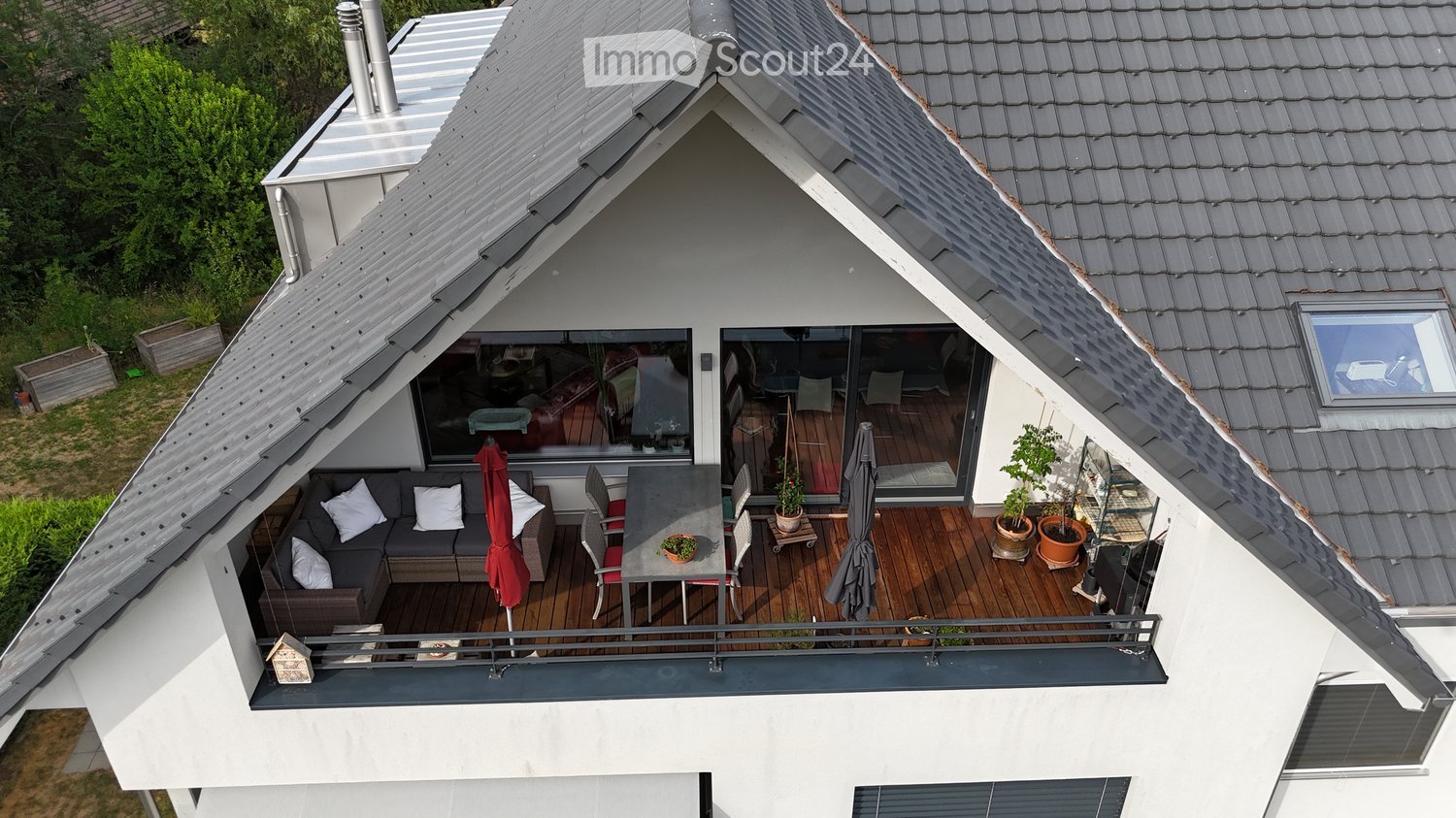 Wooden balcony with grey sofa, dining table, chairs, umbrella, and potted plants.