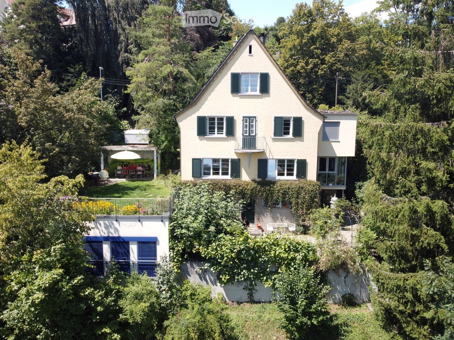 A two-story house with a yellow exterior, green shutters, and multiple windows. The house is surrounded by lush greenery and features a raised ground floor with a balcony and outdoor seating area.