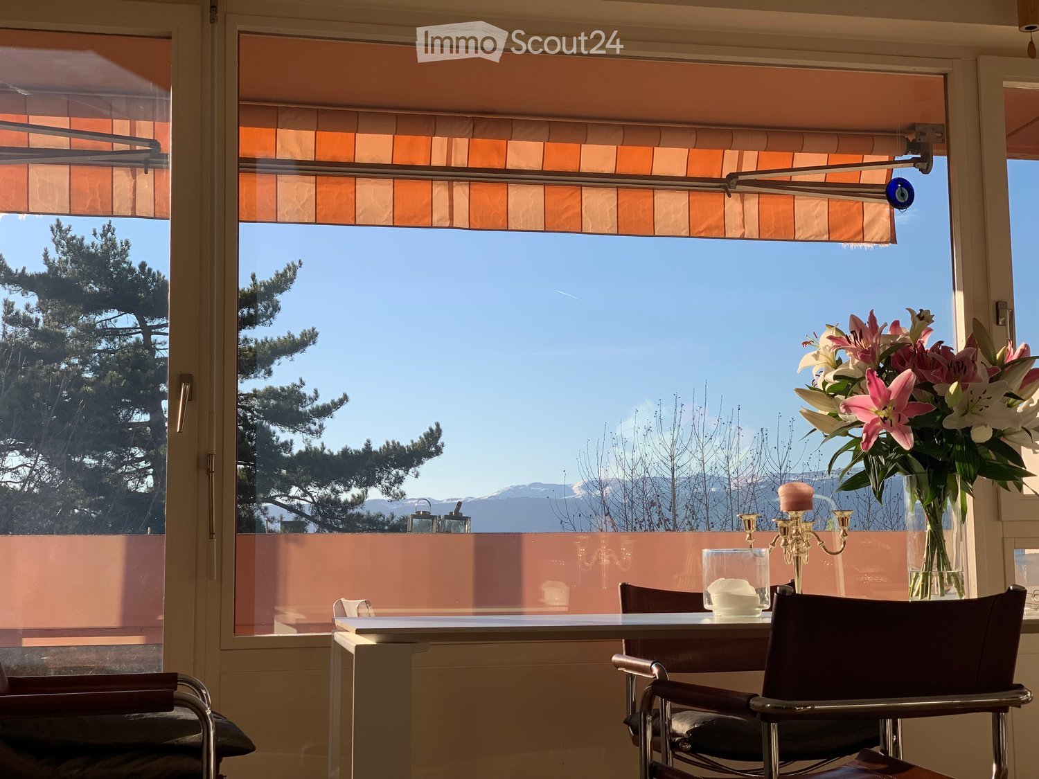 Bright room with large windows offering a scenic mountain view, furnished with a table, chairs, a vase with flowers, and a candle holder.