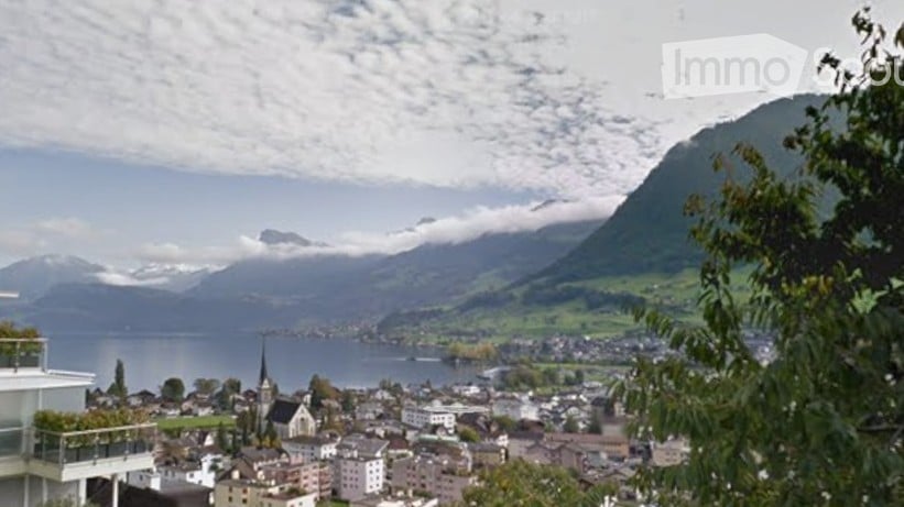 Panoramic view of a town with a lake and mountains in the background, featuring a modern balcony with greenery.