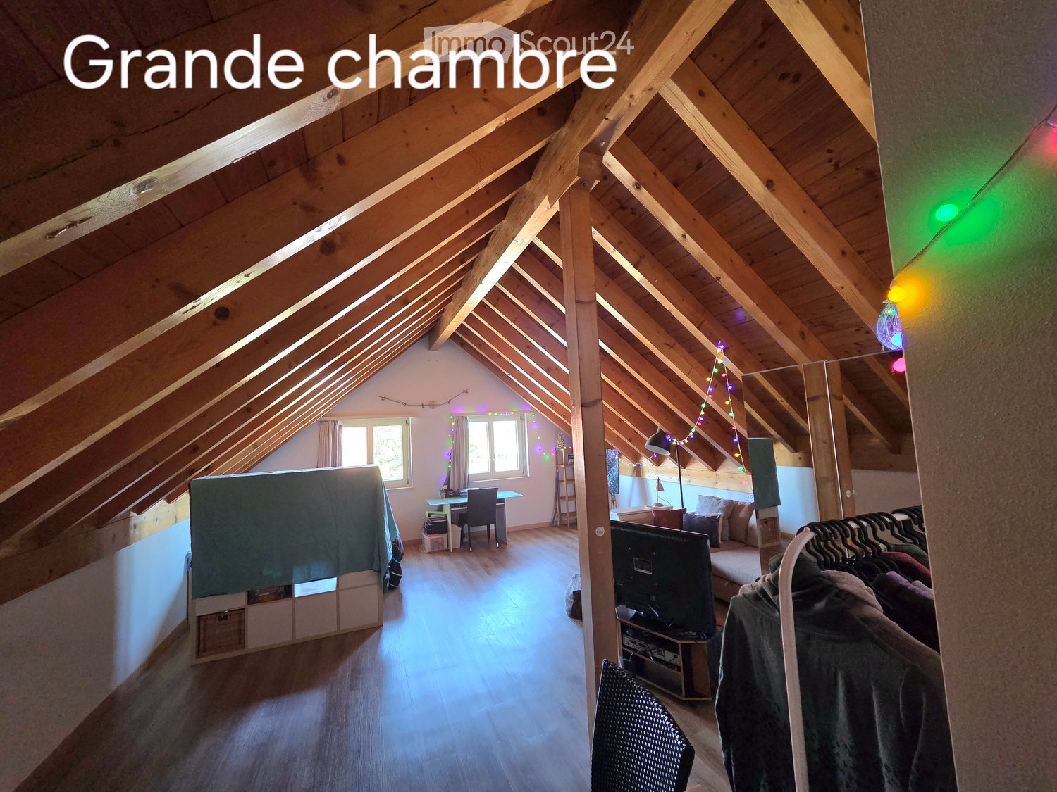 Spacious attic room with wooden beams, skylights, and large windows. Features a sofa, TV, chair, table, and wardrobe.