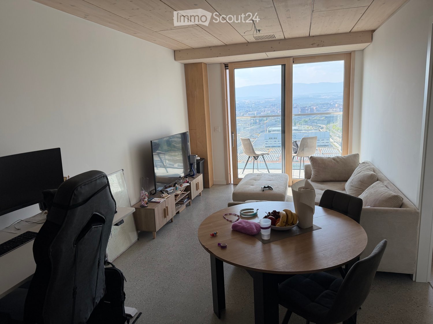 Modern living room with a view, featuring a sofa, chair, round table, laptop, screen, and a stool near the balcony.