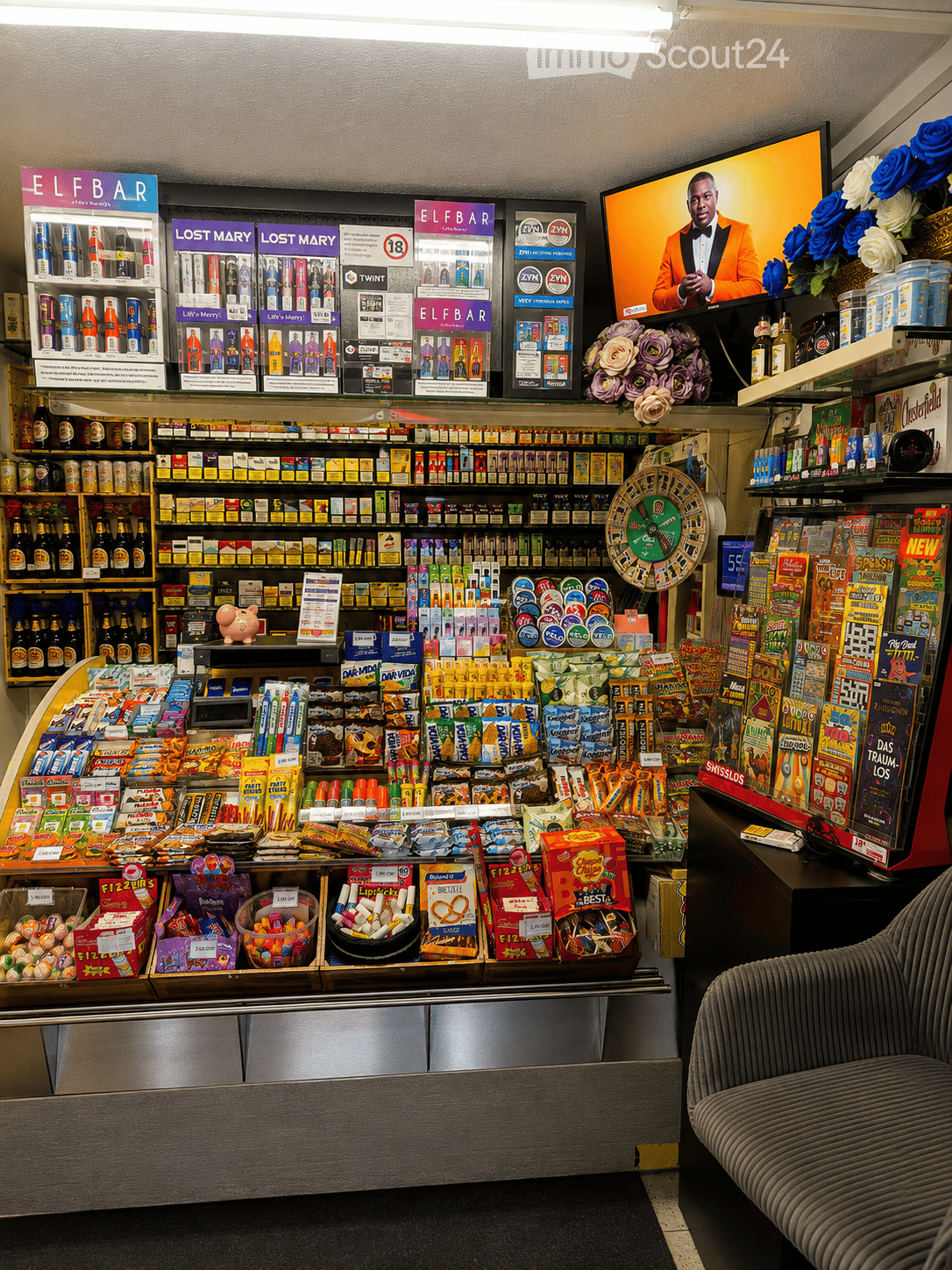 Convenience store interior with shelves stocked with various products, a television displaying a show, a sofa, and a flower arrangement on display.