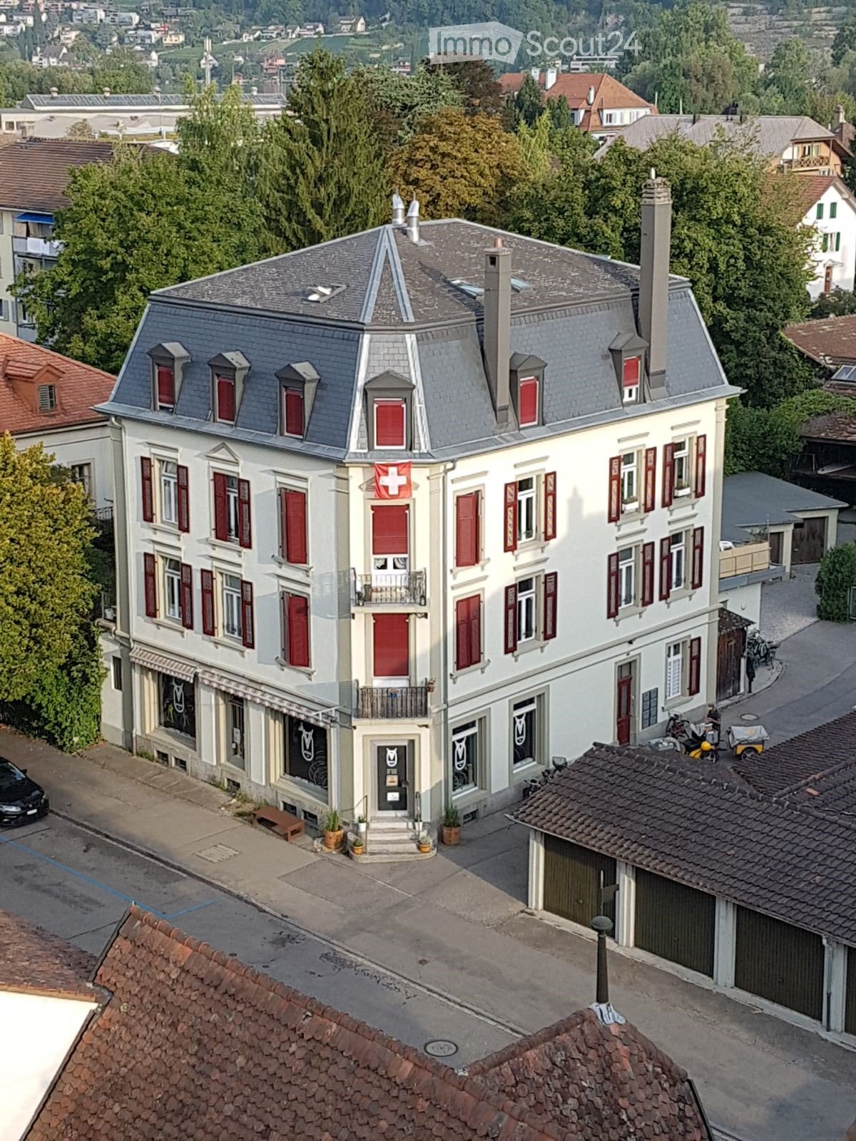 A multi-story building with a grey roof, red shutters, and a Swiss flag, surrounded by greenery and other structures.