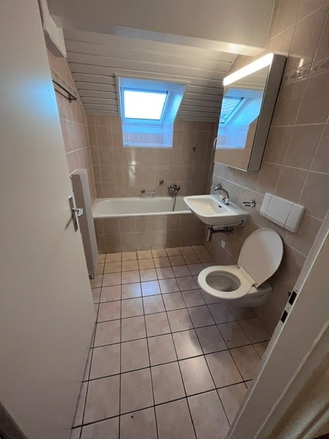 Bathroom with tiled walls and floor, bathtub, sink, toilet, and a mirror above the sink. A small window is visible above the bathtub.