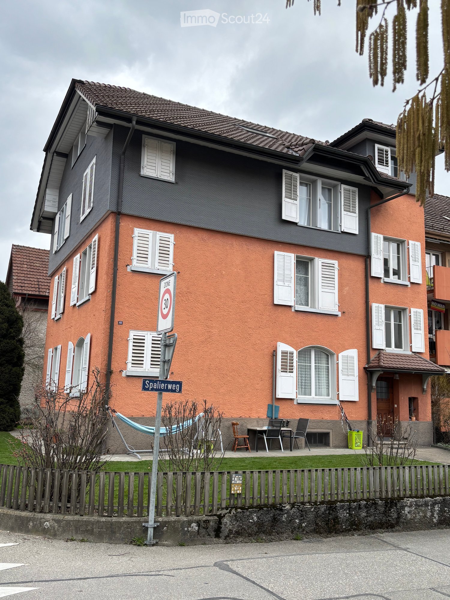 Two-story house with orange and grey exterior, multiple windows with white shutters, small garden area with patio furniture, and a speed limit sign.