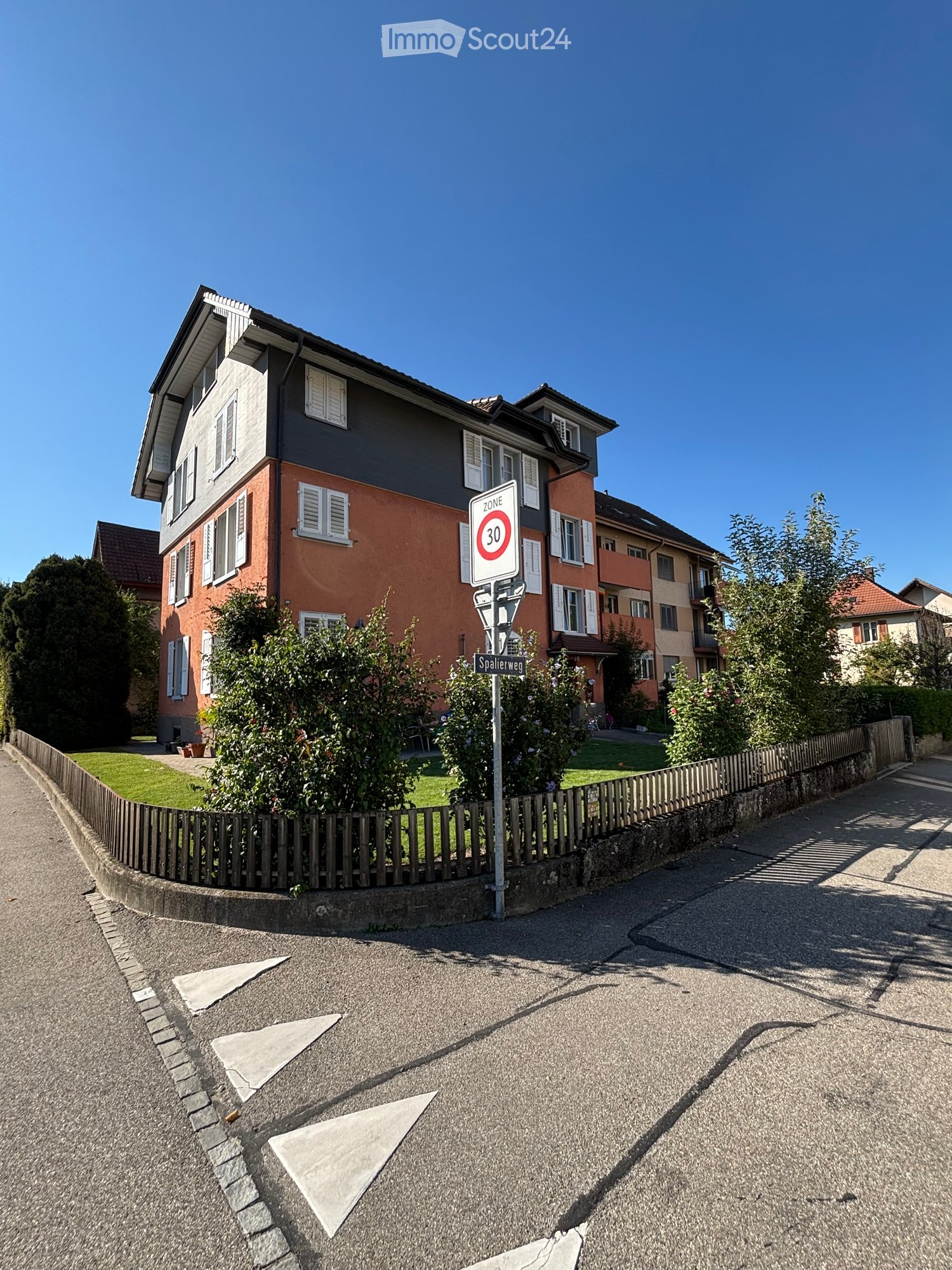 Two-story residential house with a combination of red brick and grey siding, multiple windows with white shutters, and a small garden area behind a wooden fence.