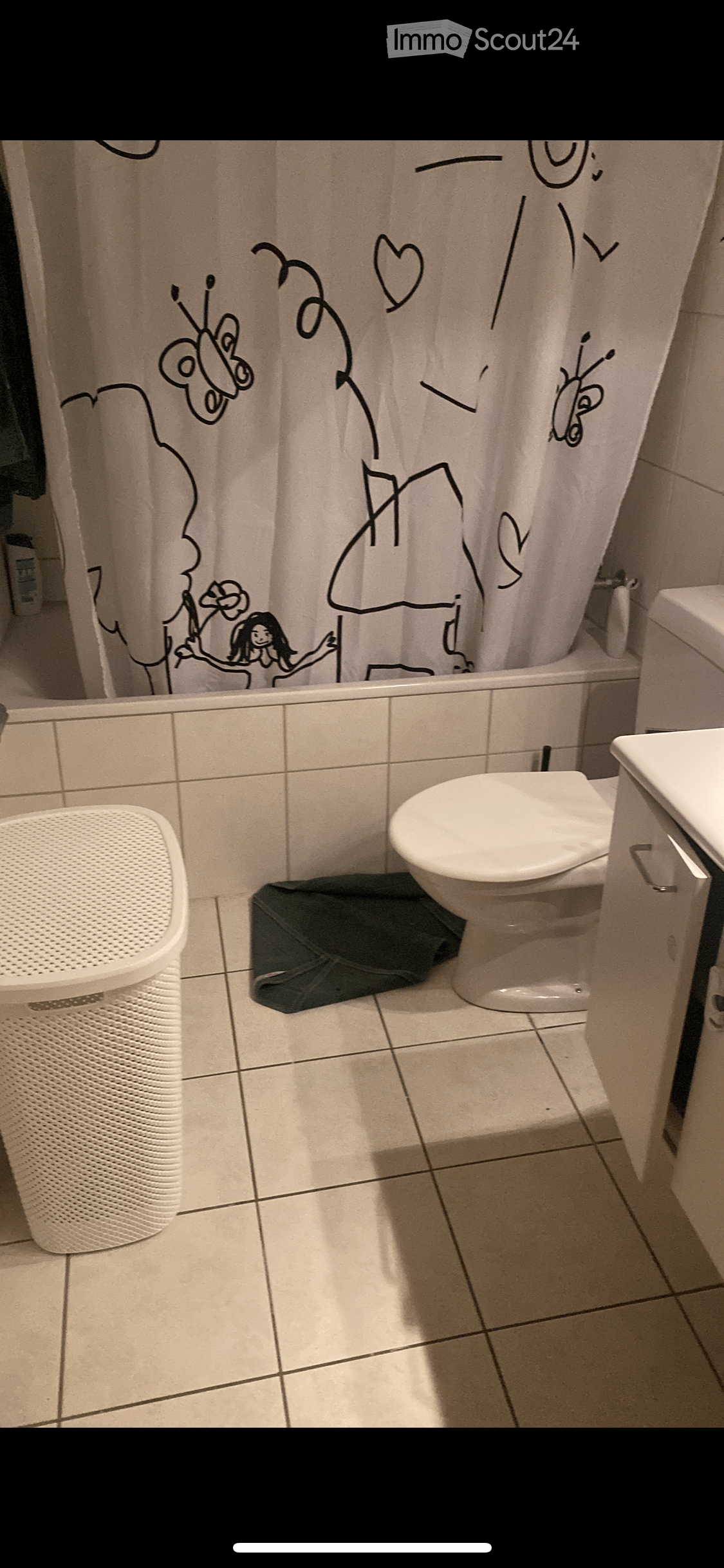 Bathroom with a toilet, sink, shower curtain featuring cartoon characters, and a white trash can. The floor is tiled, and a dark-colored bag is placed near the toilet.