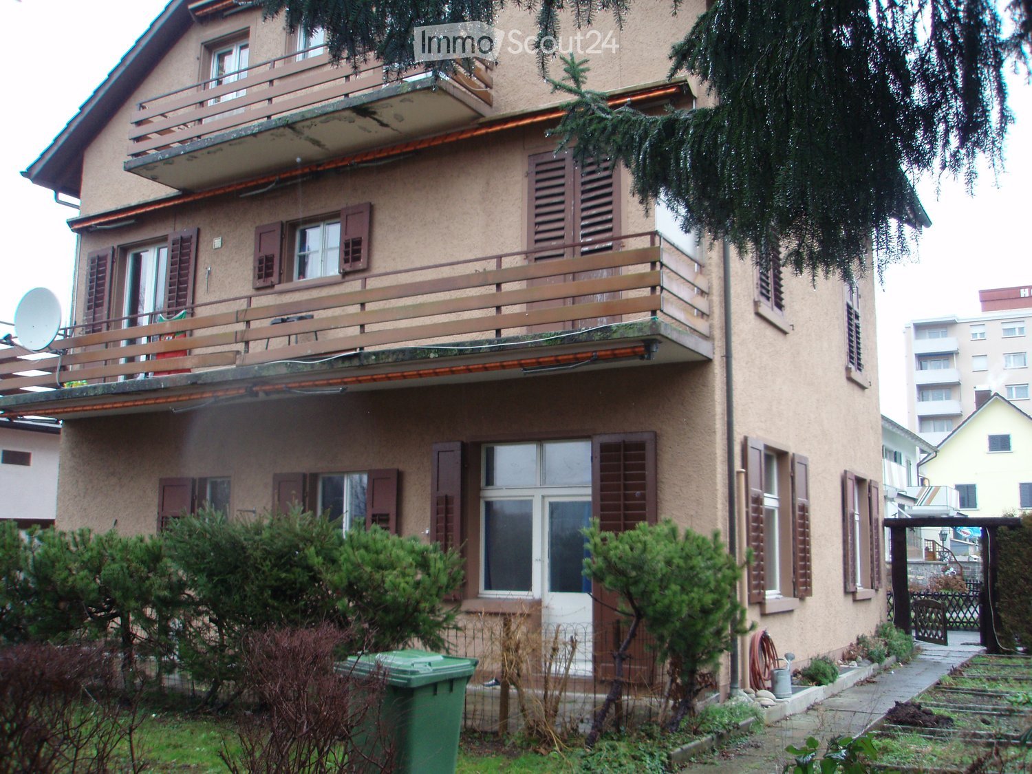 Two-story house with beige facade, multiple windows with shutters, balconies on the upper floor, and a small garden area with bushes and a green trash bin.