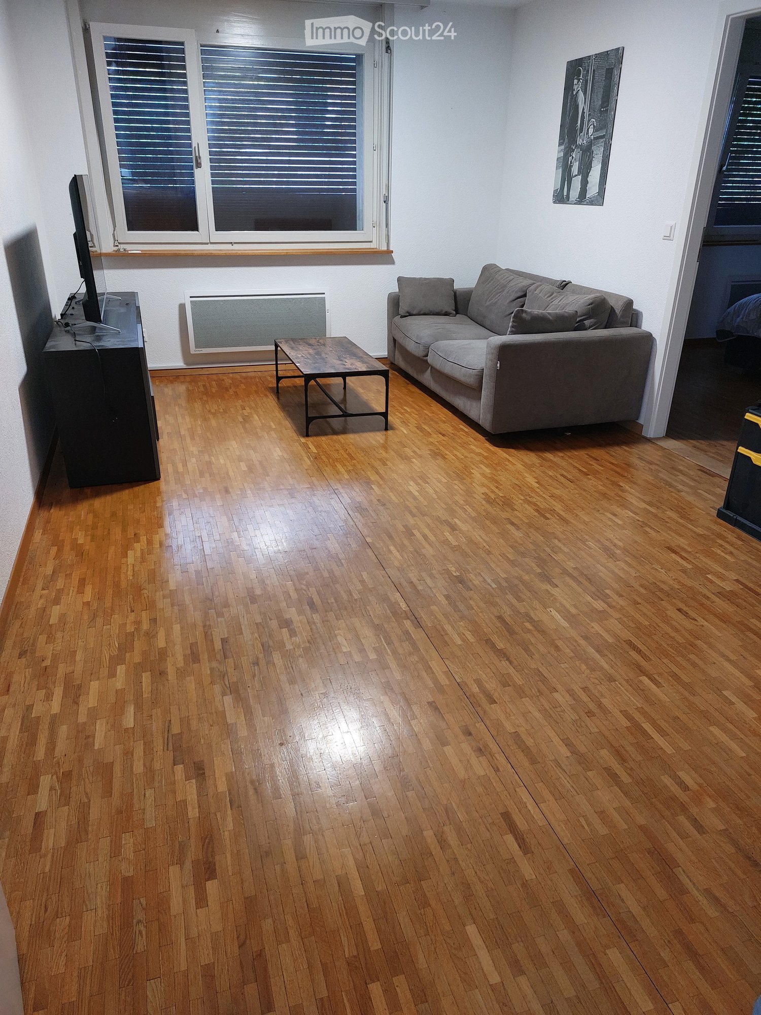 Living room with wooden flooring, grey sectional sofa, coffee table, TV stand, and large windows with blinds.