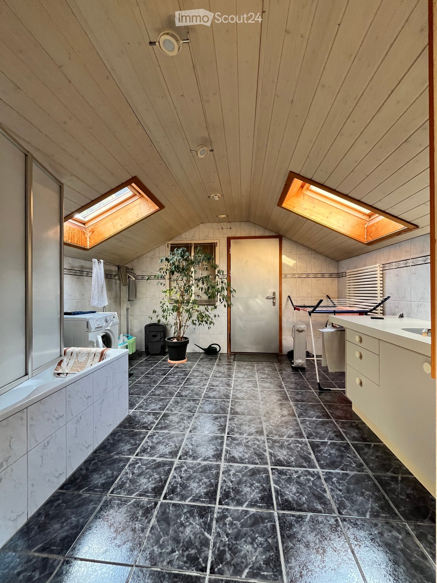 A well-lit attic space with dark tiled flooring, a bathtub, a washing machine, a sink, a potted plant, and a door leading to another room.
