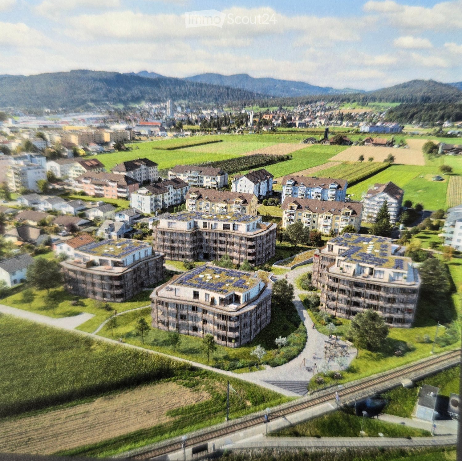 Aerial view of a residential complex with multiple apartment buildings, surrounded by green spaces, pathways, and a train track running alongside.