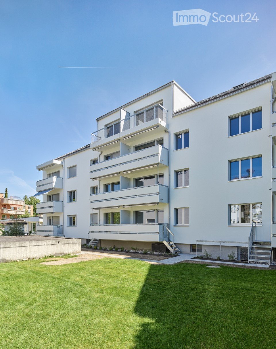 Modern multi-story apartment building with white facade, multiple balconies, and a well-maintained lawn in front.