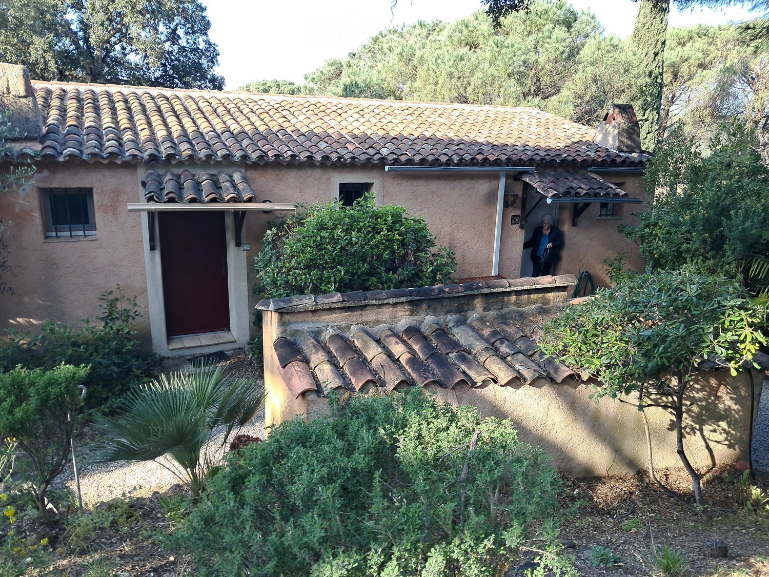 A small, rustic house with a tiled roof and light pink walls, surrounded by lush greenery and a stone wall.