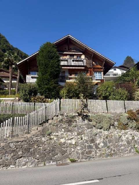 A two-story wooden house with balconies, surrounded by greenery and a stone wall.