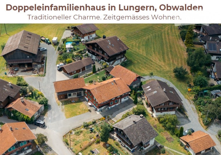Aerial view of a residential area with multiple traditional houses featuring pitched roofs, surrounded by green fields and trees.