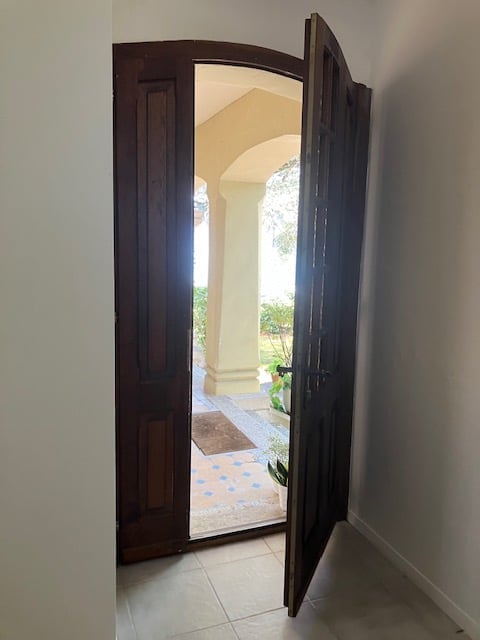 Open wooden double doors leading to a bright outdoor area with a tiled floor and visible greenery beyond.