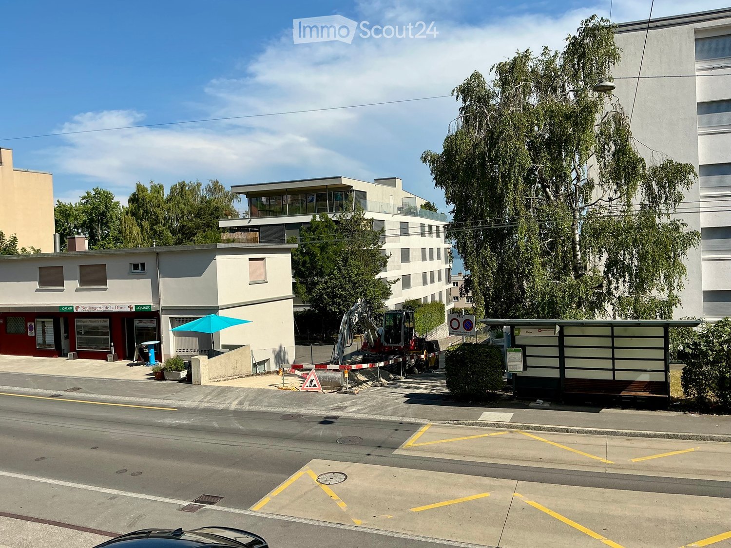 A street view showing a mix of buildings, including a small commercial building with a red facade and a modern multi-story building with large windows and balconies. There is a construction area with machinery, a bus stop, and a tree-lined sidewalk.