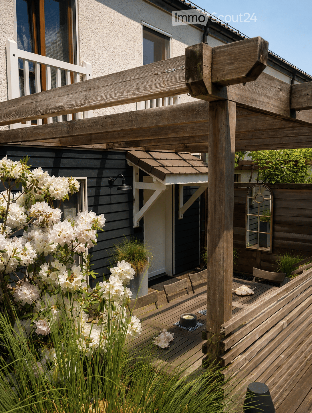 Wooden patio with a table, chairs, and a small candle, surrounded by white flowering shrubs and greenery, with a wooden pergola and a modern house in the background.