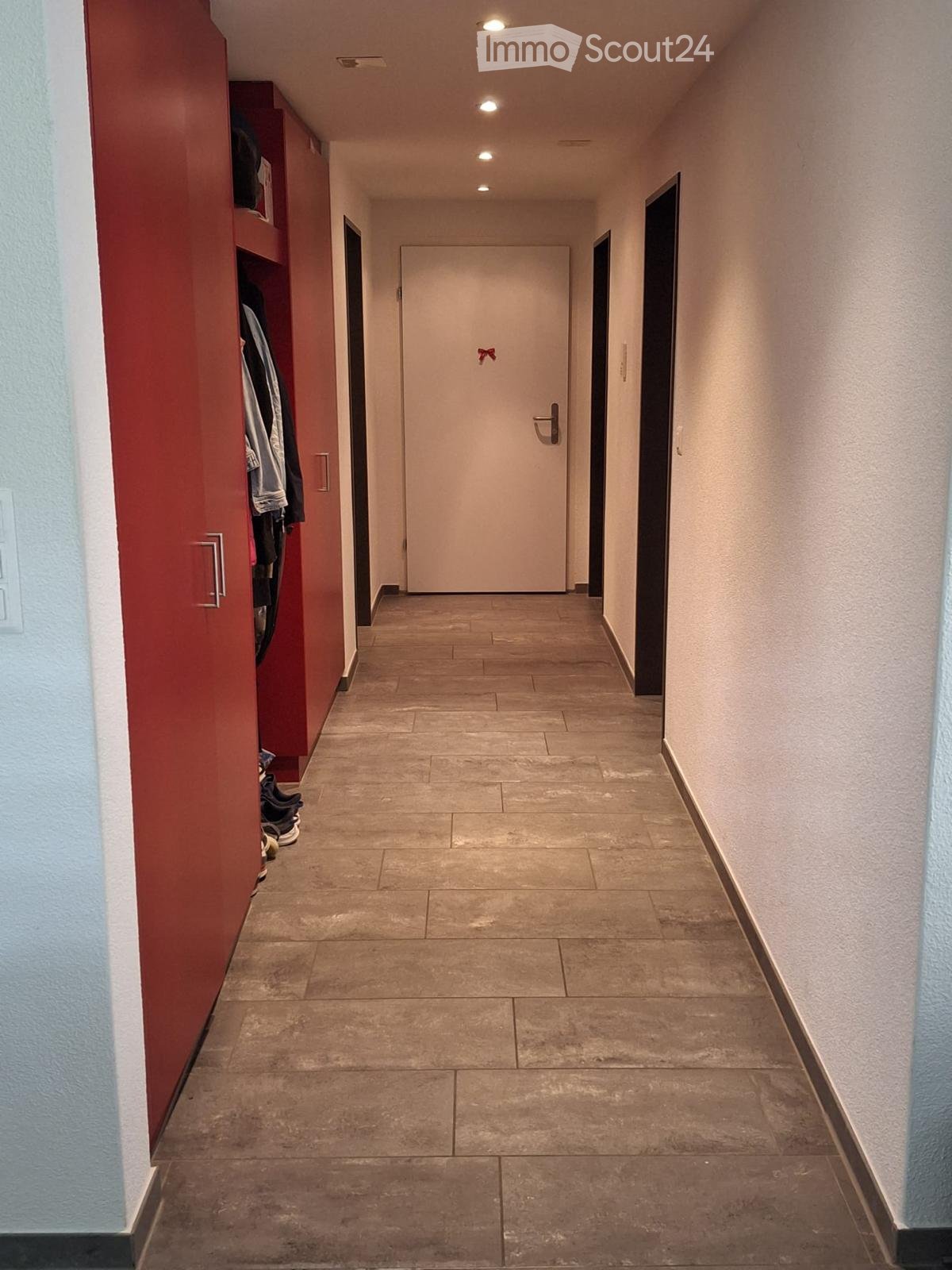 Clean hallway with tiled floor, beige walls, and a closed door at the end. A red wardrobe with hanging clothes is visible on the left side.