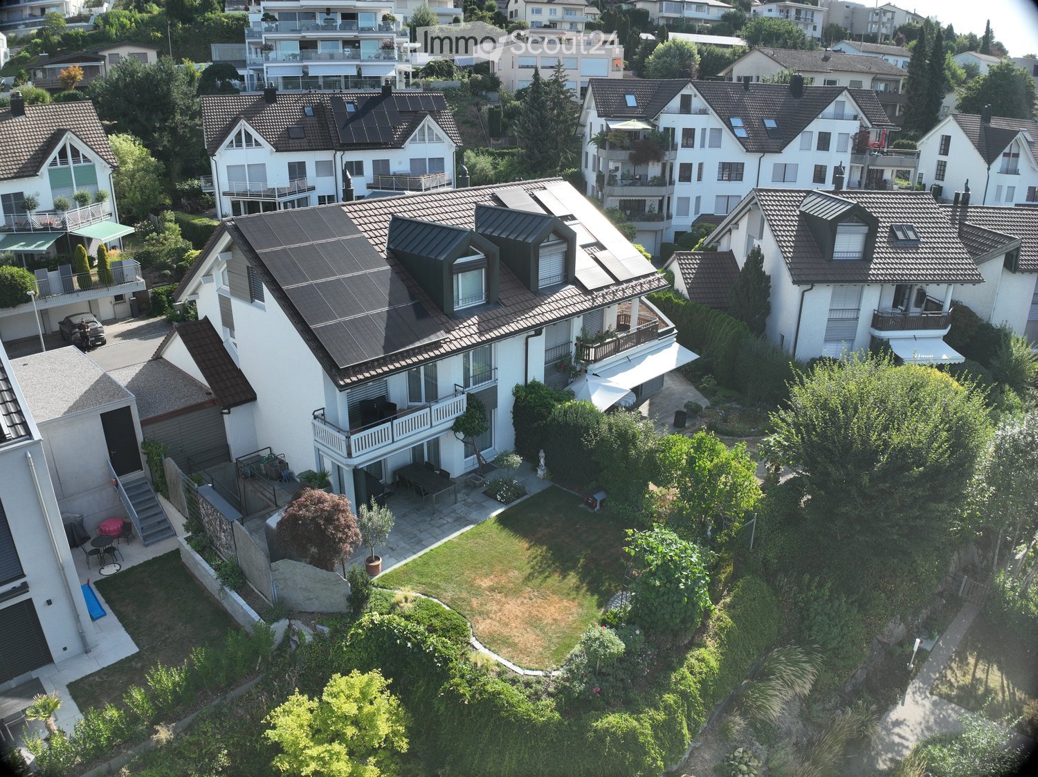 Aerial view of a modern two-story house with a well-maintained garden, featuring a patio, outdoor furniture, and surrounding greenery.