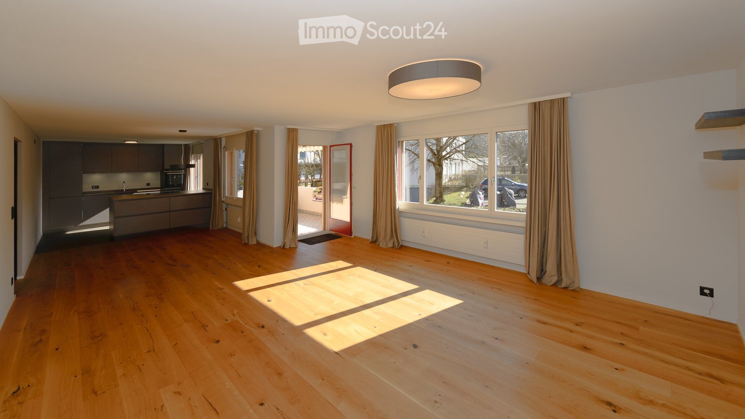 Open-plan living and kitchen area with wooden flooring, modern kitchen cabinets, and large windows with curtains.