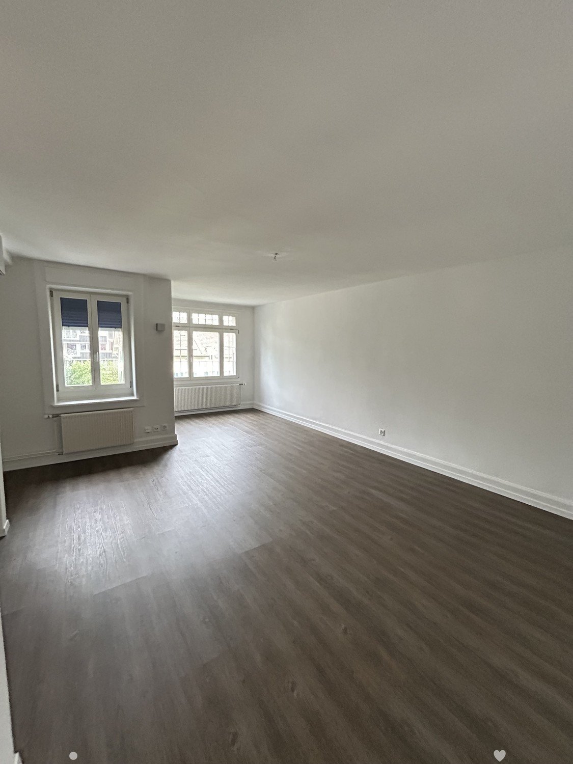 Empty room with wooden flooring, white walls, and large windows with radiators below them.