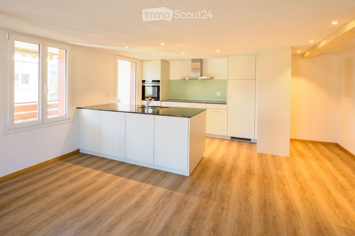 Modern kitchen with white cabinets, a central island, and a built-in oven. The space features wooden flooring and large windows.
