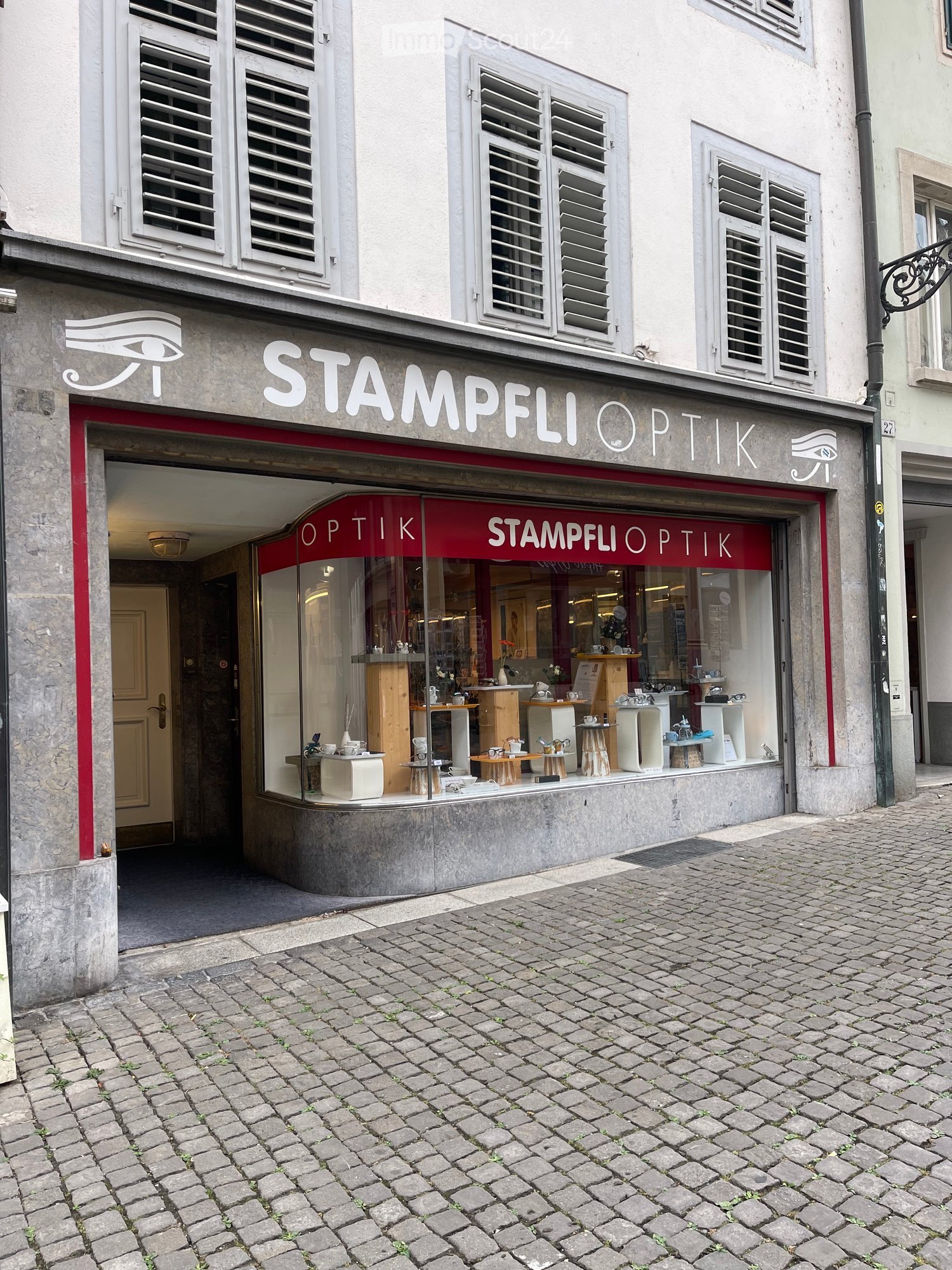 Facade of a shop named "Stampfli Optik" with a glass display window showing various optical items, located on a cobblestone street.