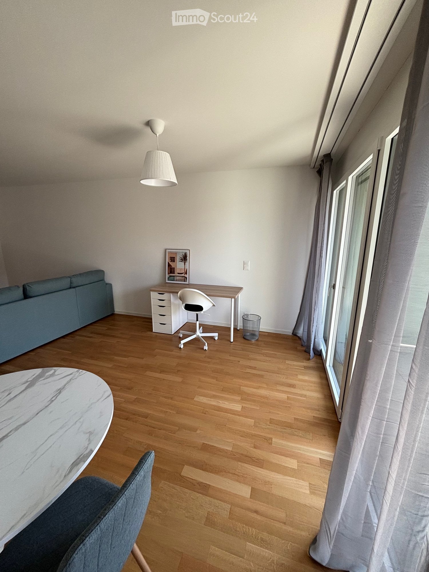 A modern, well-lit room with wooden flooring, a grey sofa, a white desk with a chair, a round marble table, and a hanging lamp.