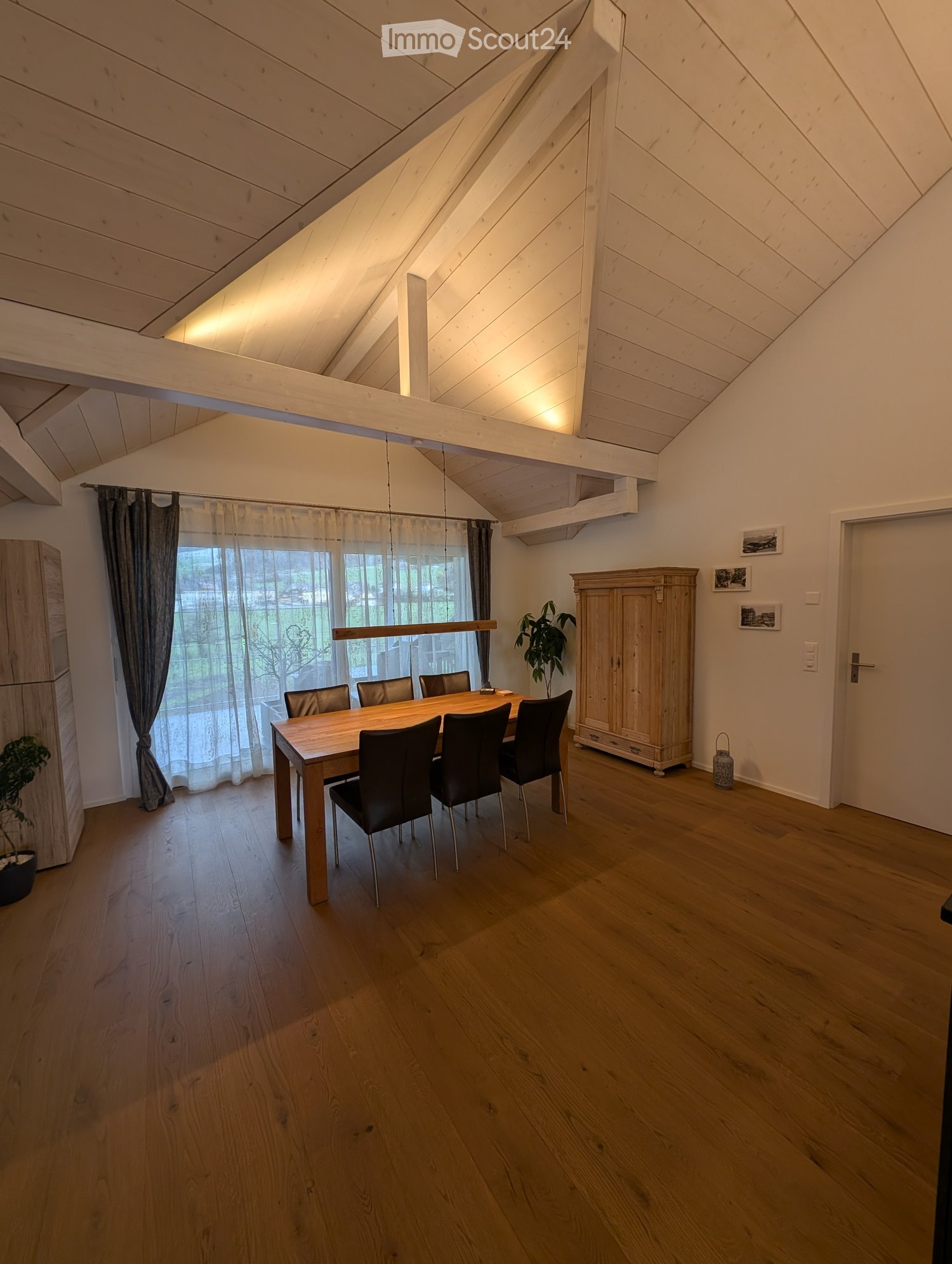 Bright dining room with wooden floors, a large wooden dining table surrounded by black chairs, a tall wooden cabinet, and large windows with curtains.