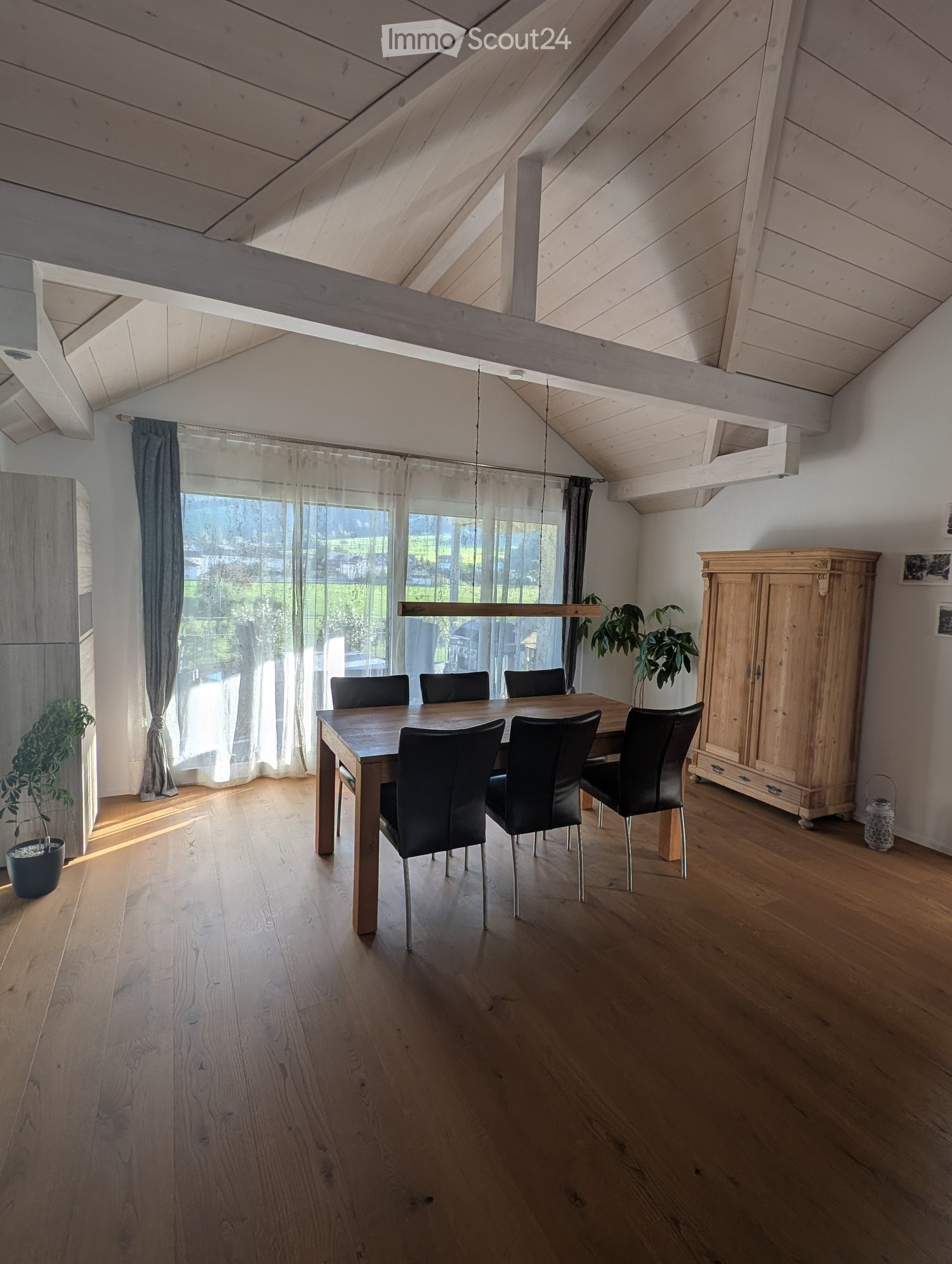 Bright dining room with wooden floor, large window with sheer curtains, dining table with six black chairs, wooden cabinet, and potted plants.
