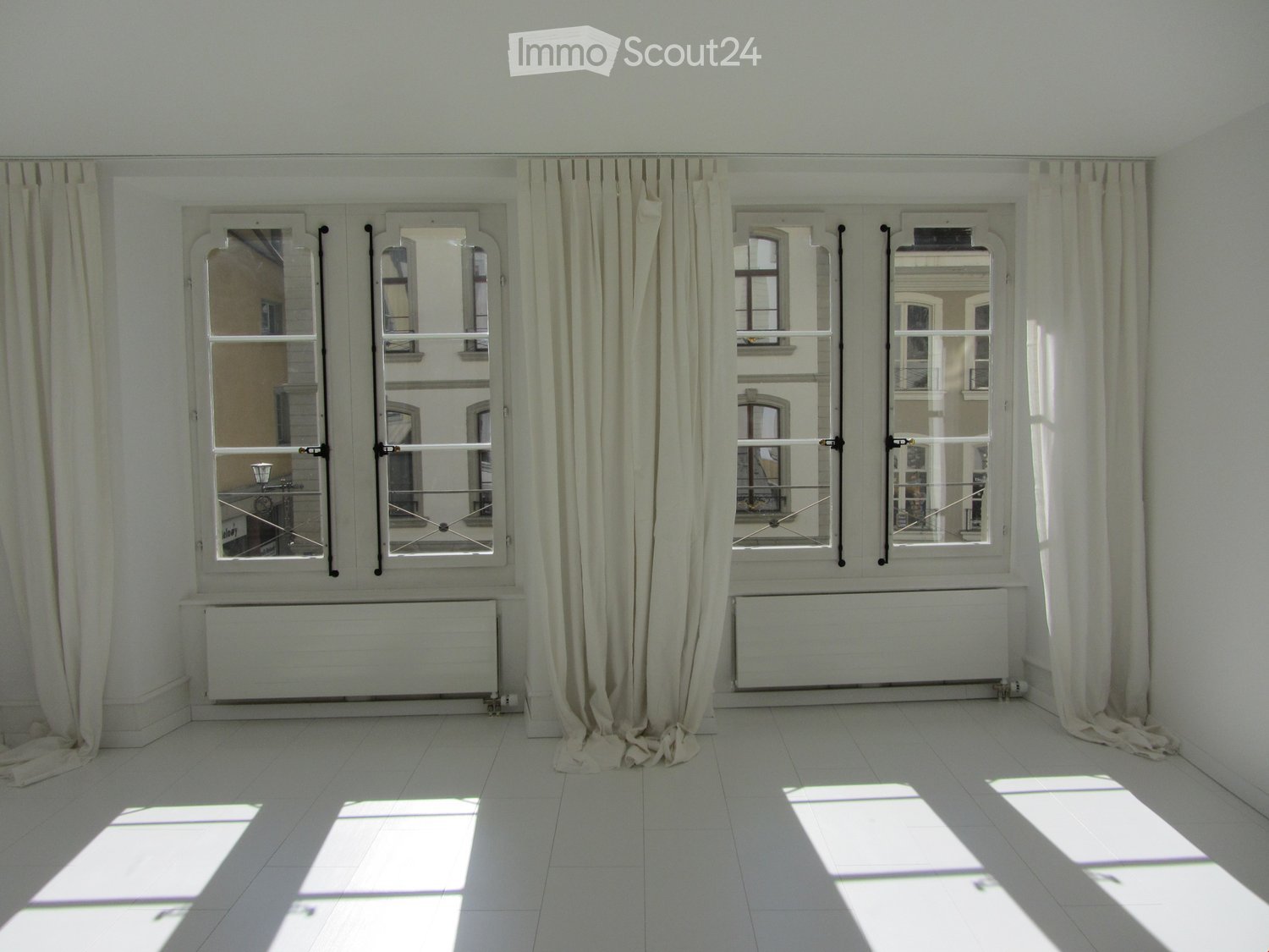Empty room with white tiled floor, large windows with beige curtains, radiators below the windows, and natural light streaming in.