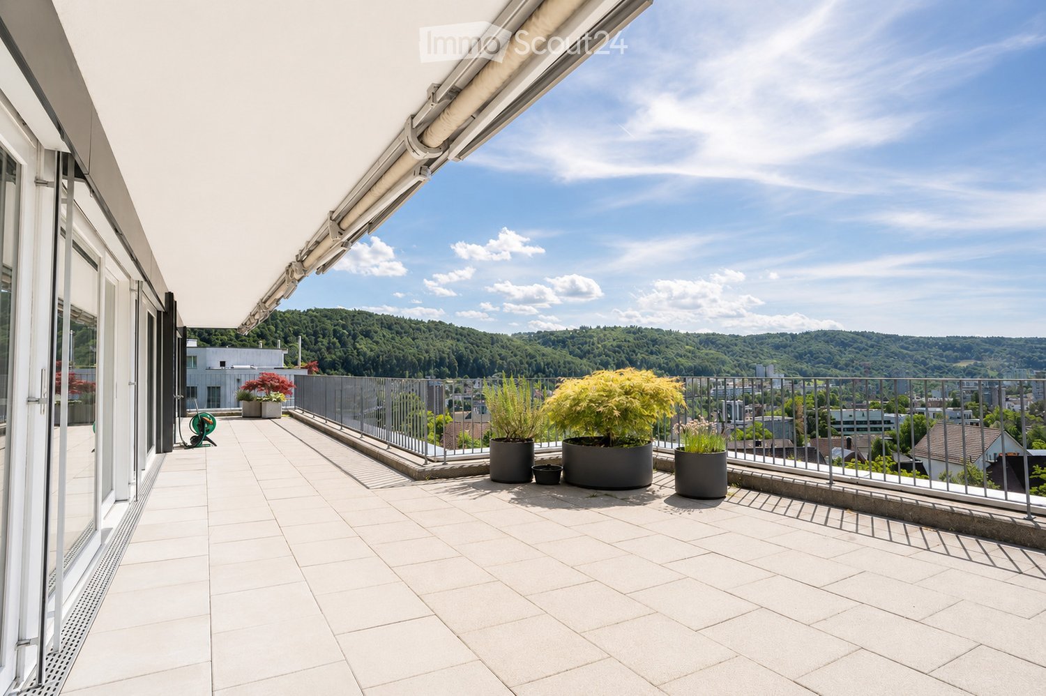 Balcony with white tiled floor, metal railing, potted plants, and a scenic view of a green, hilly landscape.