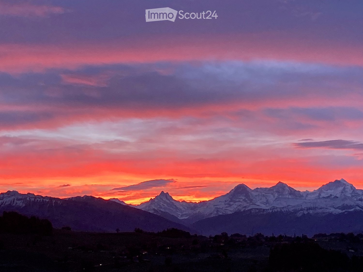 A scenic view of snow-capped mountains at sunset, with a vibrant sky of pink and orange hues, and a small settlement visible in the foreground.
