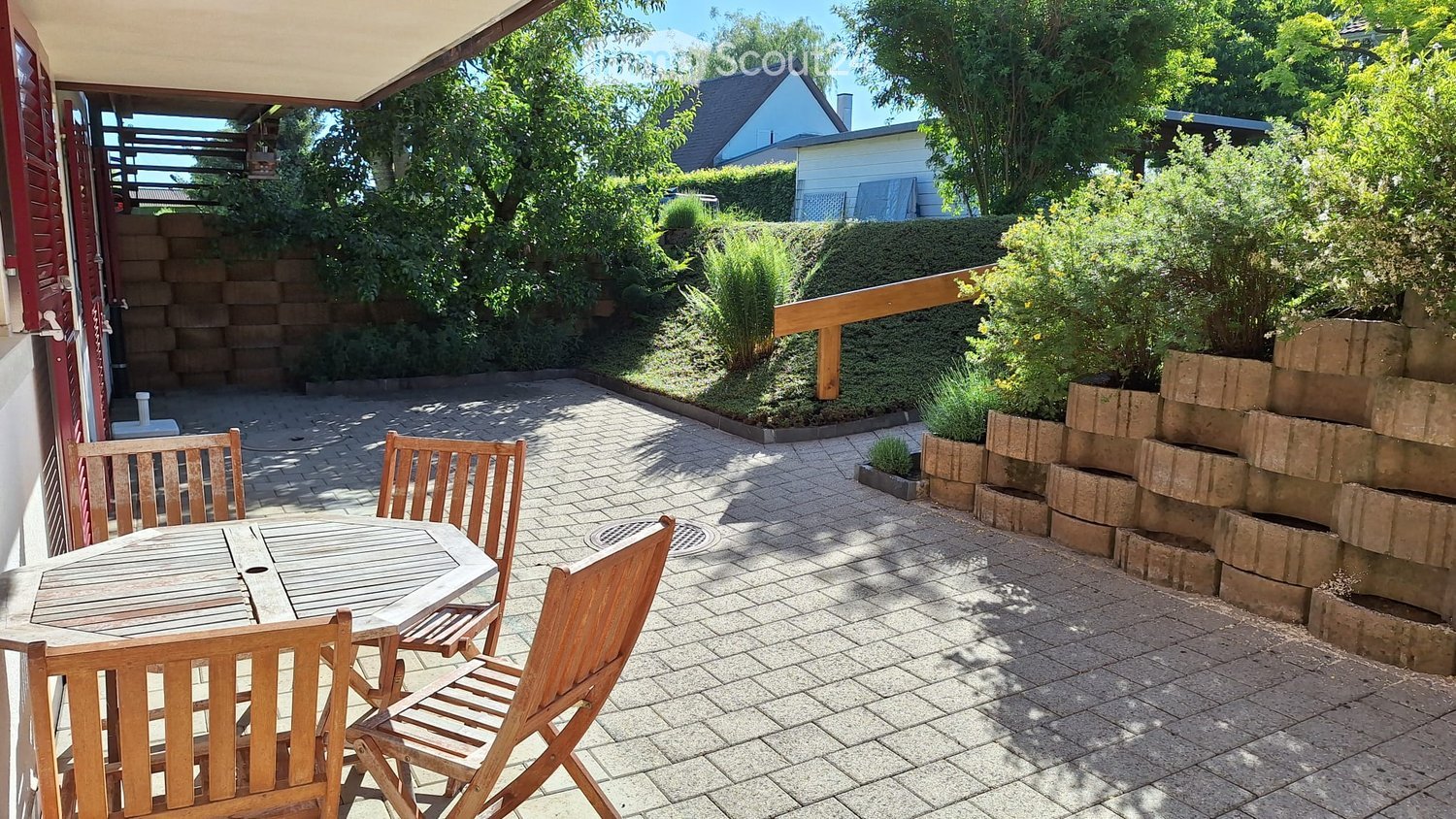 A sunny patio with a wooden table and four chairs, surrounded by greenery and a brick wall.