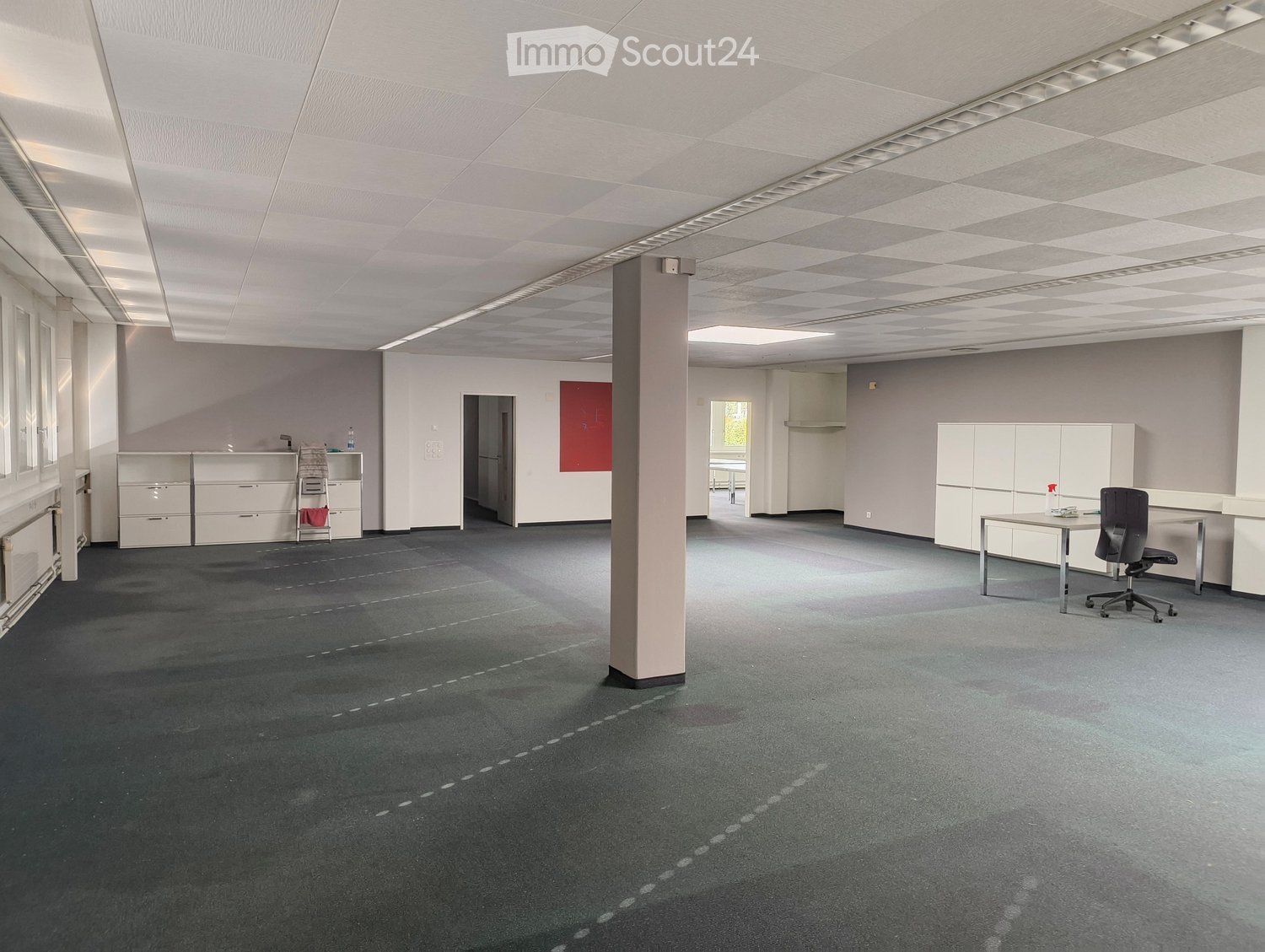 Office space with grey carpet, white walls, ceiling lights, a central pillar, and minimal furniture including a chair, table, and cabinets.