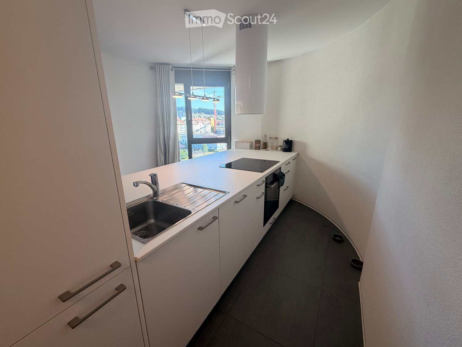Modern kitchen with white cabinets, stainless steel sink, induction cooktop, and built-in oven. Features a large window with city view and pendant lighting.