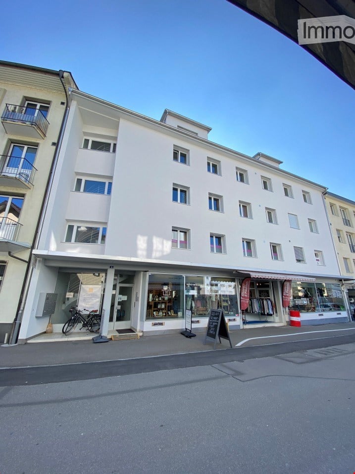 Modern multi-story building with white facade, large windows, and balconies. Ground floor features commercial storefronts with glass windows and signage.